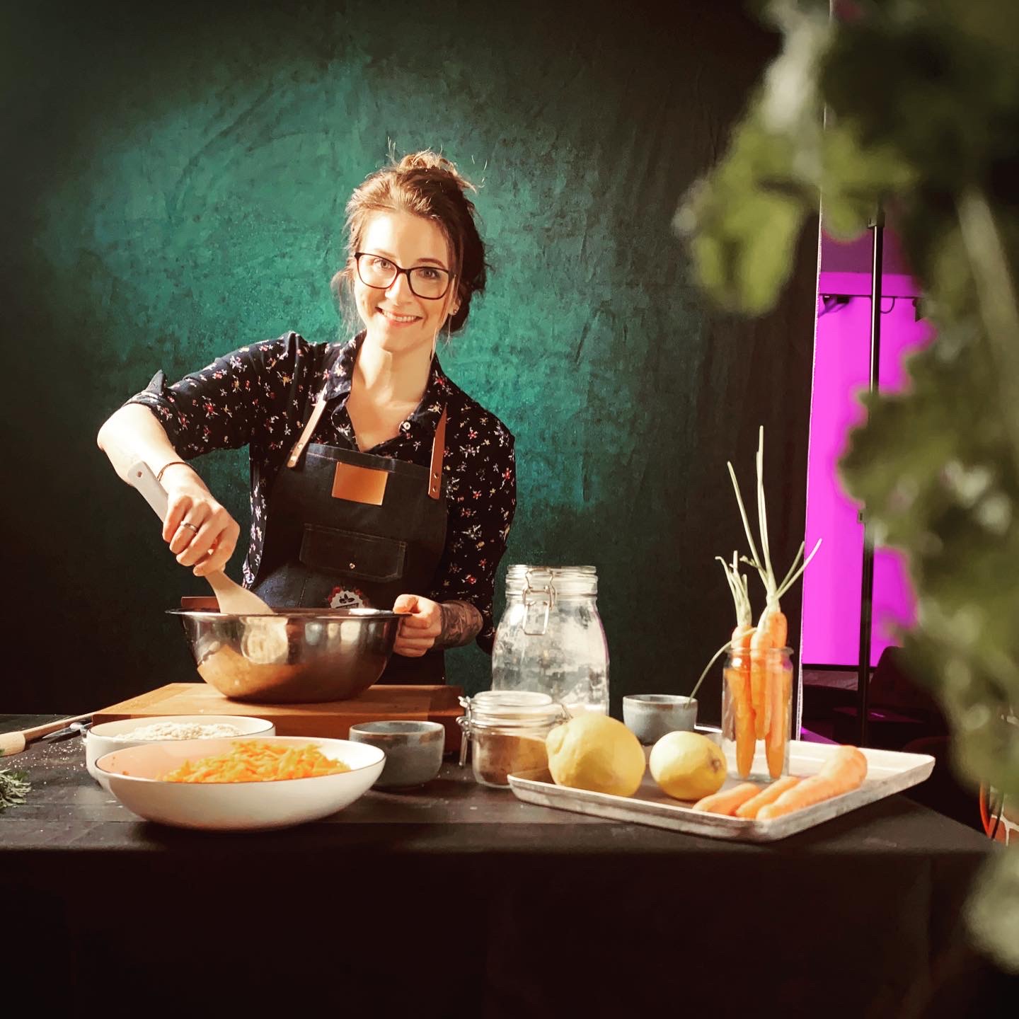 Eine Frau in Schürze rührt in einer Schüssel auf einem Tisch mit Karotten, Zitronen und Gläsern, vor grünem Hintergrund.
