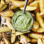 Ein Glas grüner Dip mit Löffel, umgeben von knusprigen Kartoffelspalten und Rosmarin.