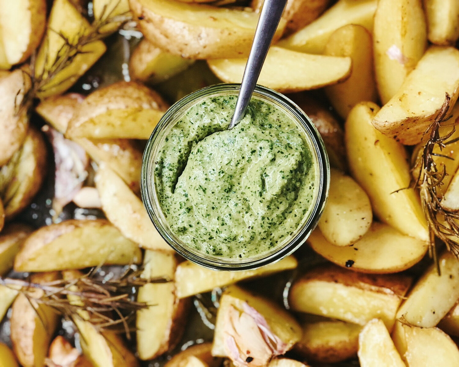Ein Glas grüner Dip mit Löffel, umgeben von knusprigen Kartoffelspalten und Rosmarin.