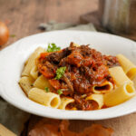 Rigatoni mit Tomatenfleischsauce, garniert mit Petersilie, auf einem weißen Teller, im Hintergrund eine Zwiebel.