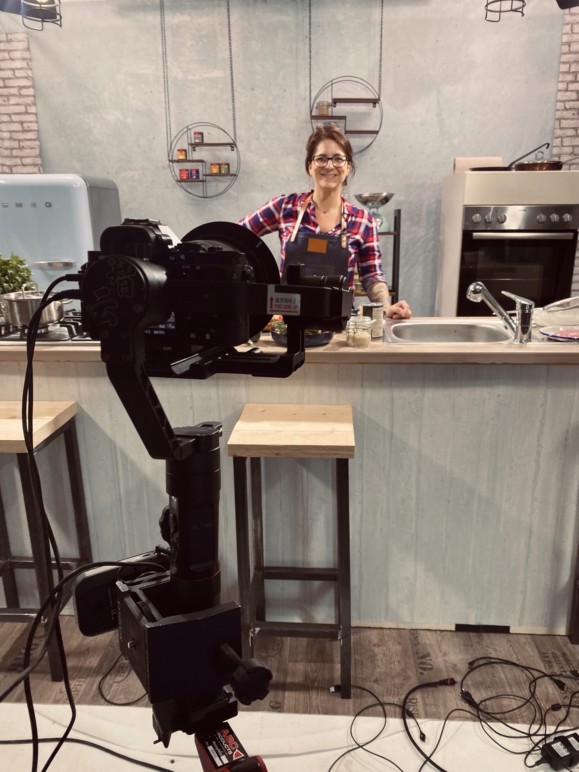Frau in Küche mit kariertem Hemd und Schürze, im Hintergrund Kamerasetup und Backofen sichtbar.