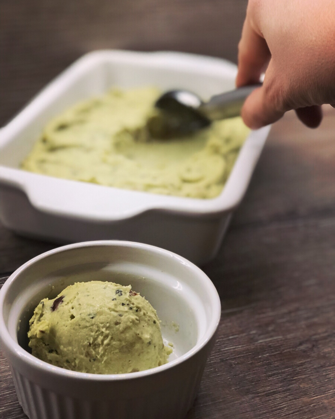 Avocado-Eis Nicecream ohne Eismaschine Hand schöpft grünes Avocado-Eis mit einem Eisportionierer aus einer weißen Form, davor eine Schale mit einer Kugel.