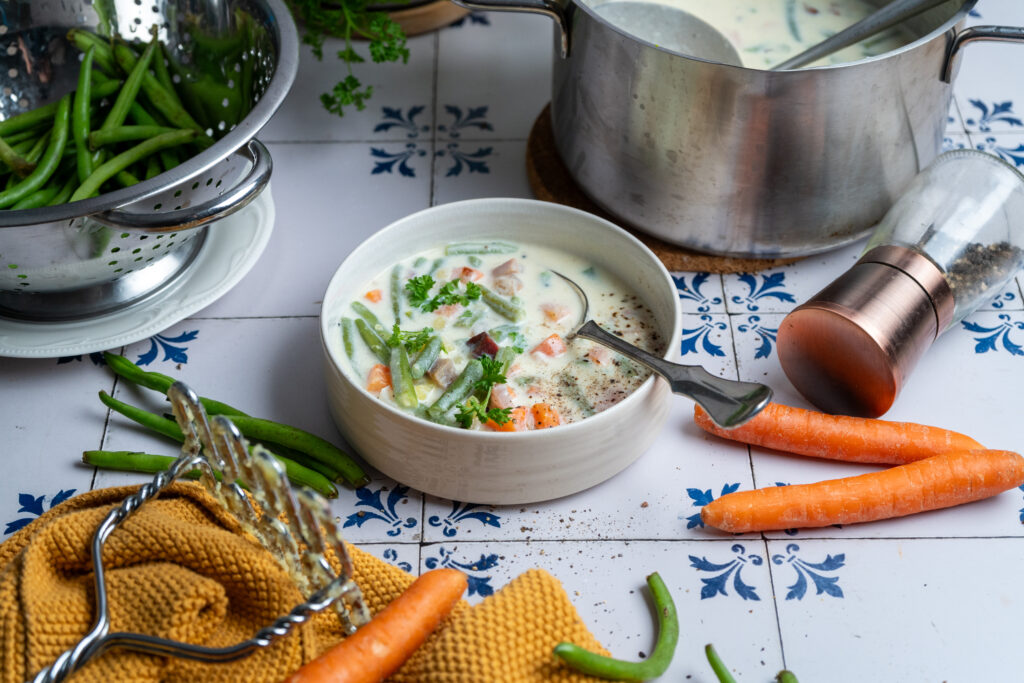 Rheinische Bohnensuppe mit Buttermilch, daneben grüne Bohnen und Möhren auf blau-weißen Fliesen