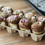 Cupcakes in Waffelbechern mit Schokoglasur und bunten Streuseln in einem Eierkarton auf Holztisch.