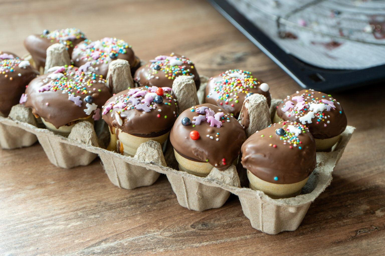 Cupcakes in Waffelbechern mit Schokoglasur und bunten Streuseln in einem Eierkarton auf Holztisch.