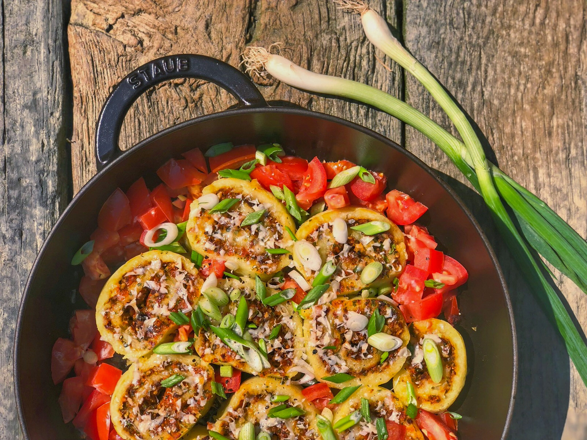 Gefüllte Zucchinischeiben mit gehackten Tomaten und Frühlingszwiebeln in einer schwarzen Pfanne auf Holzuntergrund.