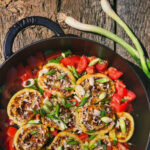 Bratpfanne mit gefüllten Teigtaschen, Tomaten und Frühlingszwiebeln auf einem Holztisch, daneben drei Frühlingszwiebeln.