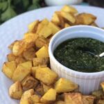 Gebackene Kartoffelwürfel mit einer Schale grüner Sauce in der Mitte, serviert auf einem weißen Teller.