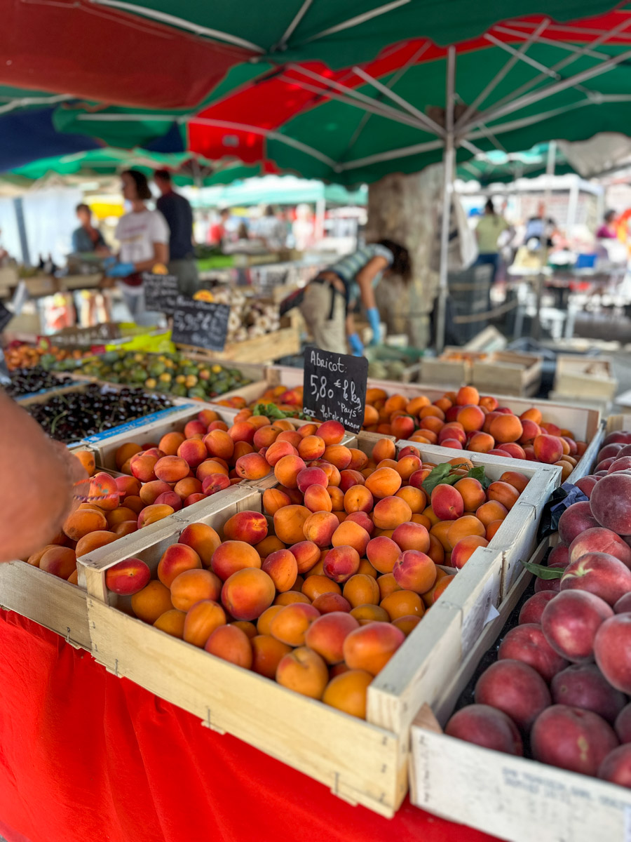 Aprikosen Crumble-3 Kisten mit Aprikosen und Pfirsichen auf einem Marktstand unter bunten Sonnenschirmen. Menschen im Hintergrund.