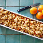 Rechteckige Form mit Aprikosenstreuselkuchen auf blauem Fliesentisch, daneben frische Aprikosen und ein Messer.