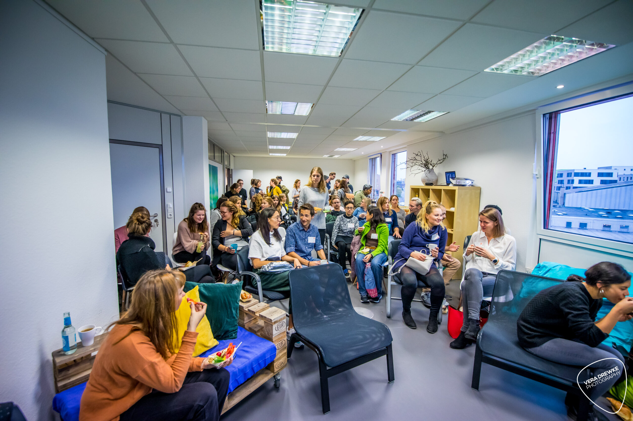 Creativ-Mornings-6 Menschen sitzen in einem Raum und unterhalten sich, einige essen und trinken. Der Raum wirkt wie ein Veranstaltungsbereich.