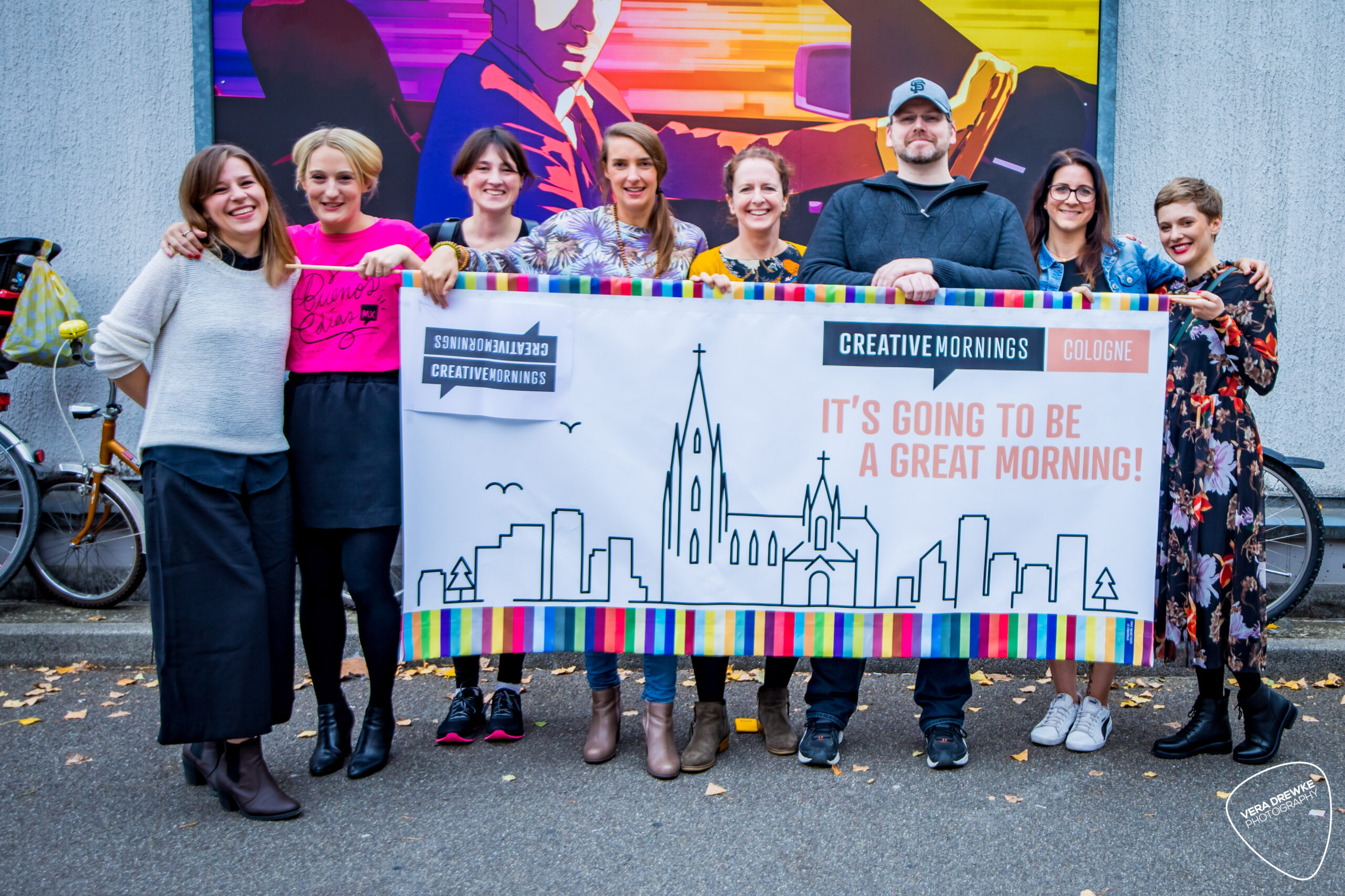 Creativ-Mornings-5 Gruppe lächelnder Menschen hält ein Banner mit Stadtsilhouette und dem Text: