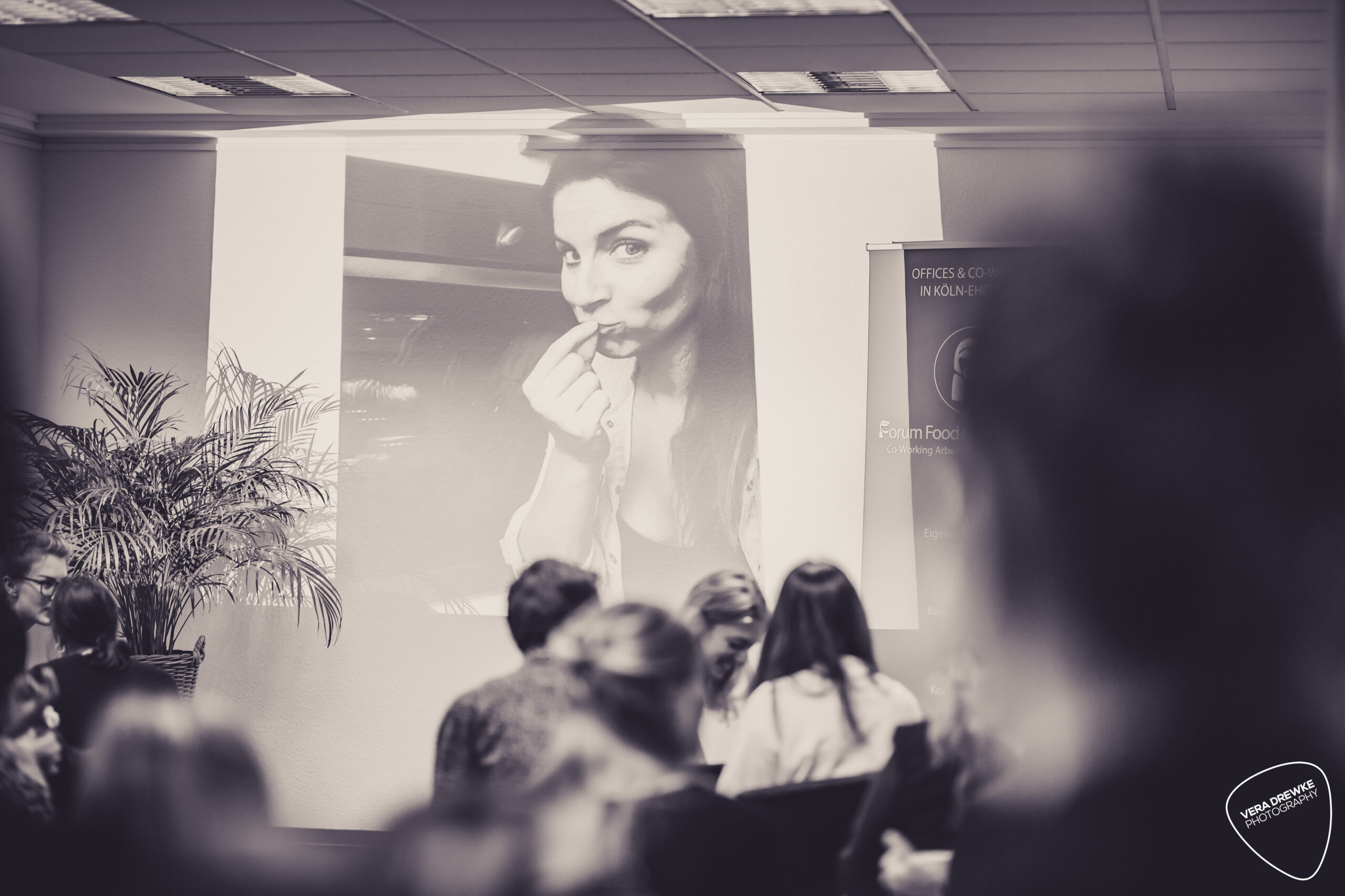 Creativ-Mornings-2 Menschen sitzen in einem Raum, ein großes schwarz-weißes Porträt einer Frau wird an die Wand projiziert.