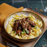 Pastaschalen mit dunklem Fleischragout und Kräutern auf einem Teller neben einem senffarbenen Stofftuch.