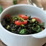 Weiße Schüssel mit Linsensalat, Kirschtomaten und Kräutern auf Holztisch. Im Hintergrund Frühlingszwiebel und Grünzeug.