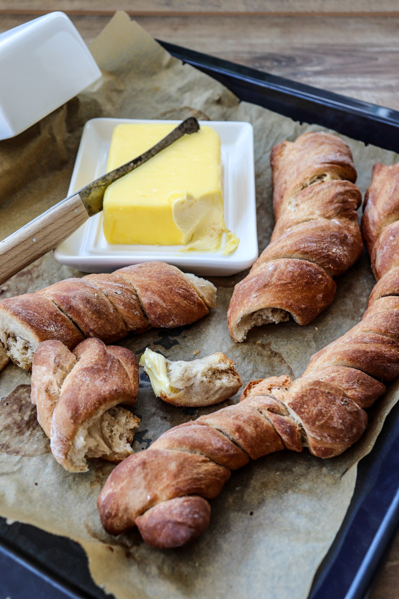 Frisch gebackene, gedrehte Brote auf Backpapier mit Butter und Messer auf einem Teller.
