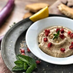 Weiße Schale mit Hummus, dekoriert mit Granatapfelkernen und Minze, auf einem Tablett mit Zitronenscheibe und Brot.