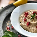 Teller mit Hummus, garniert mit Granatapfelkernen und Kräutern, auf einem Tablett mit Minzblättern und Zitronenscheibe.