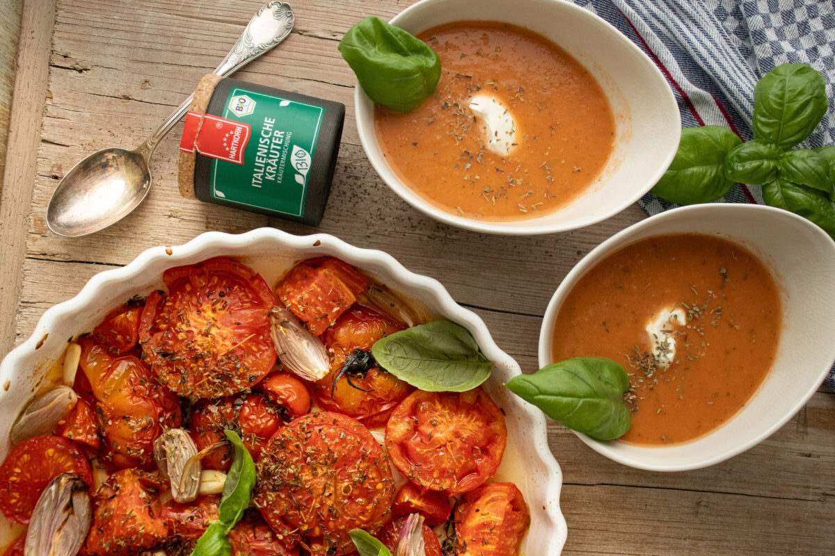 Gebackene Tomaten in einer Schale, daneben zwei Teller Tomatensuppe mit Sahneklecksen, Basilikum und Kräutermischung.