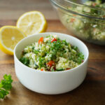 Weiße Schale mit frischem Tabouleh-Salat aus Petersilie, Couscous und Gemüse. Im Hintergrund geschnittene Zitronen.