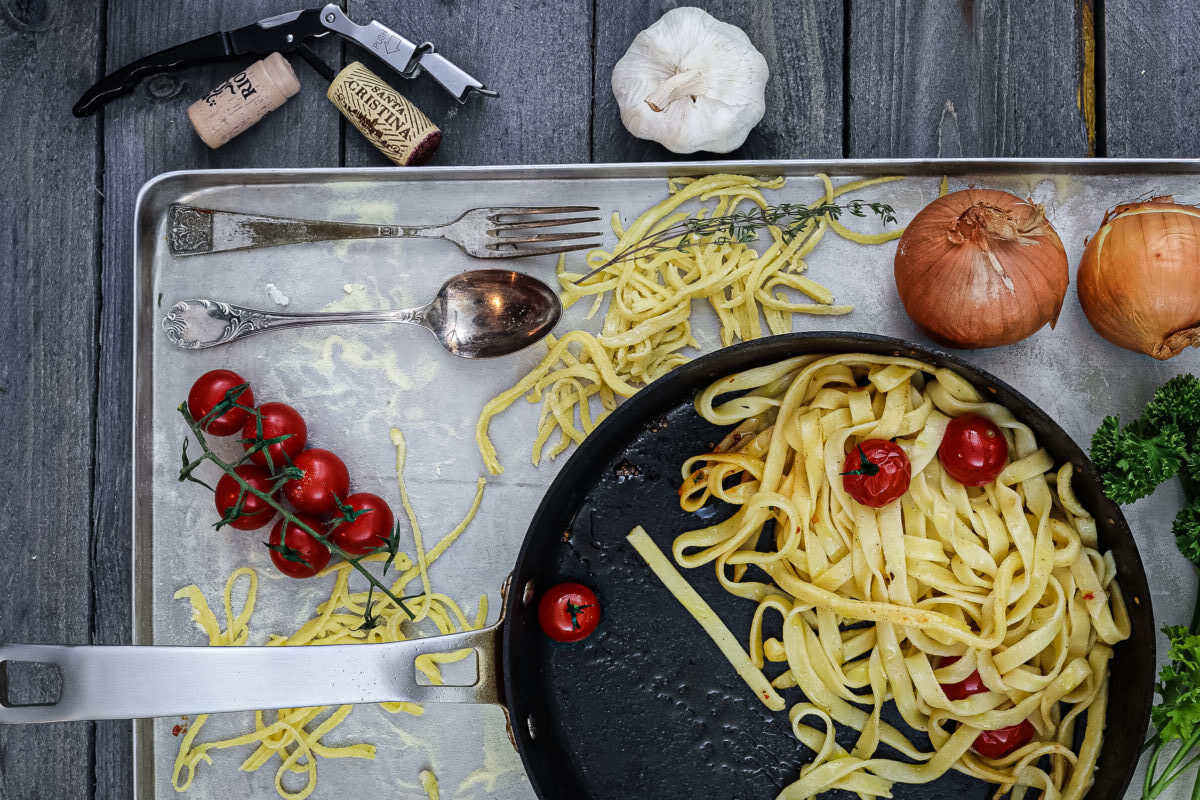 Pfanne mit Nudeln und Kirschtomaten auf Blech, daneben Knoblauch, Zwiebeln, Tomatenzweig, Löffel, Gabel und Weinöffner.