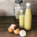 Zwei Flaschen mit gelber Flüssigkeit neben einer Flasche Likör und leeren Eierschalen auf einem Holztisch.