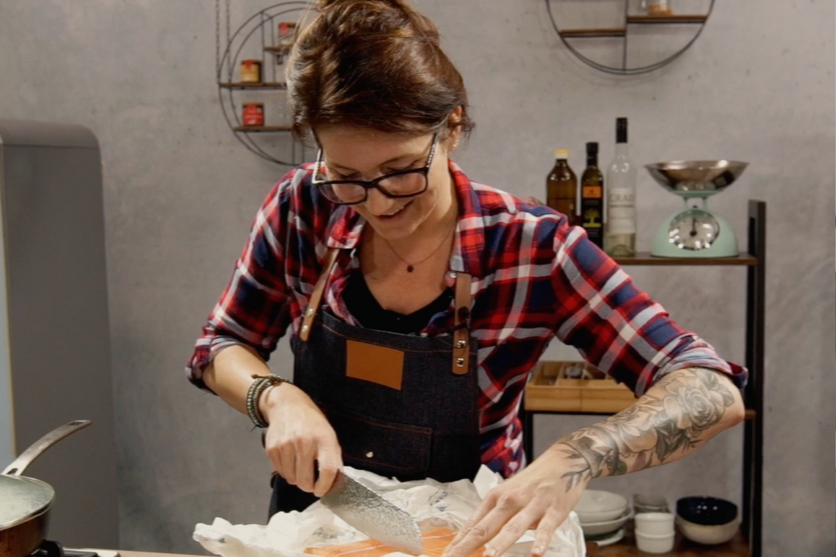 Presenterin-für-1000-Gute-Gründe-1 Frau in karierter Bluse schneidet Lachs in einer modernen Küche auf einem Schneidebrett.