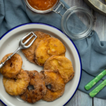 Teller mit frittierten Apfelscheiben, Gabel und Messer, daneben Apfel, Zimt und Zucker in Holzschale auf Holztisch.