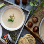 Cremige Maronensuppe mit grünen Pfefferkörnern, Knoblauch, Brot, Kastanien und Kräutern auf einem Tisch angerichtet.