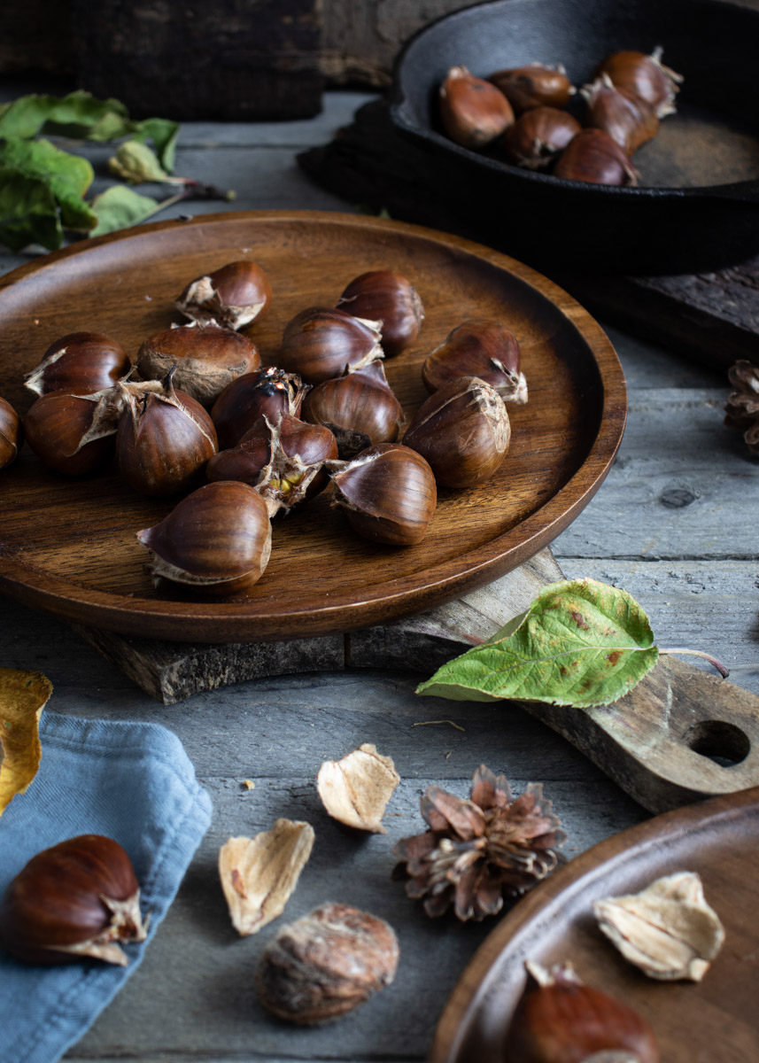 Holzteller mit Kastanien auf rustikalem Tisch, umgeben von Blättern und Schalenstücken.