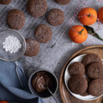 Schokoladenkekse auf einem Gitter und Teller, mit Puderzucker bestäubt; daneben Mandarinen und geschmolzene Schokolade.