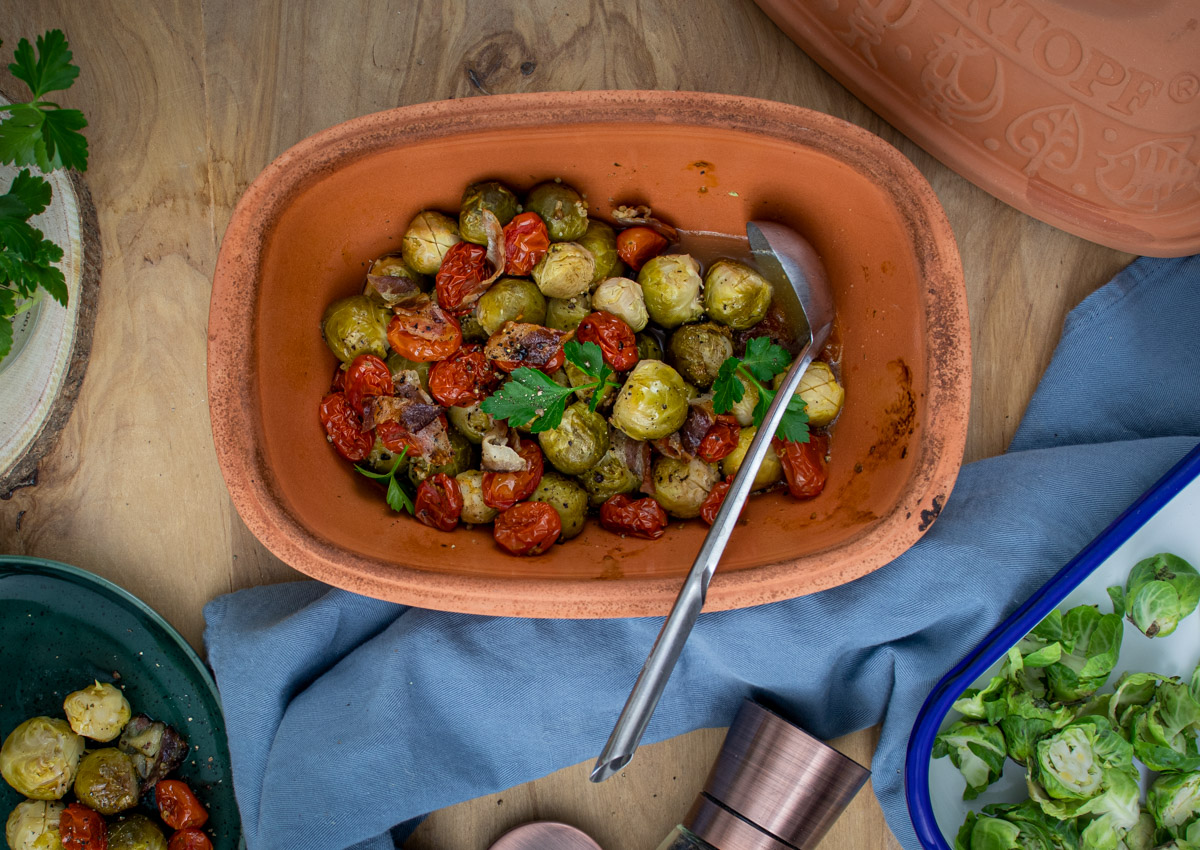 Rosenkohl_aus_dem_römertopf_3 Gebratener Rosenkohl und Kirschtomaten in einem Tontopf, serviert mit einem Löffel und garniert mit Petersilie.