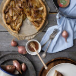 Zwiebel-Tarte auf Holztisch; ein Stück fehlt, daneben Gabeln, Honig, Schalotten und eine Springform auf Serviette.