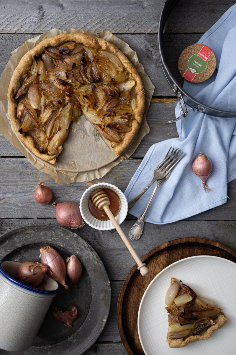 Zwiebel-Tarte auf Holztisch; ein Stück fehlt, daneben Gabeln, Honig, Schalotten und eine Springform auf Serviette.