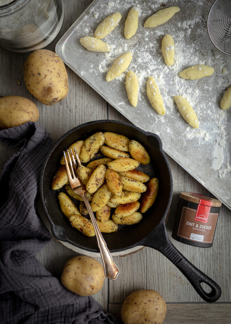 Pfanne mit gebratenen Schupfnudeln, daneben rohe Schupfnudeln auf Blech, Kartoffeln und Zimt-Zucker-Gewürz.