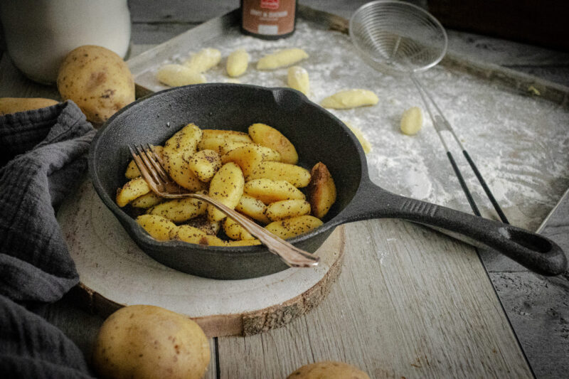 Bratkartoffeln in einer gusseisernen Pfanne mit Gabel auf Holzbrett, umgeben von rohen Kartoffeln und Küchentuch.