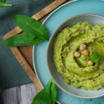 Grüner Hummus in einer Schale mit Kichererbsen, flankiert von Brotscheiben und frischen Blättern auf Holzbrett.