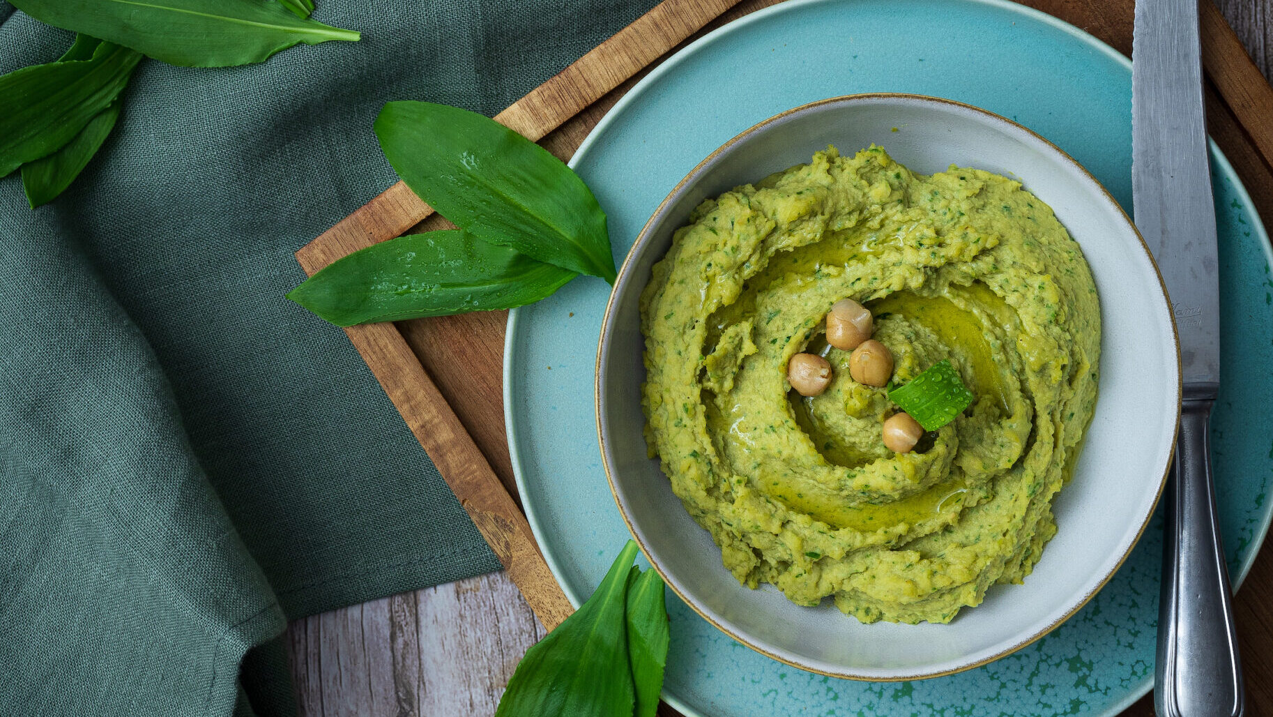 Grüner Hummus in einer Schale mit Kichererbsen, flankiert von Brotscheiben und frischen Blättern auf Holzbrett.