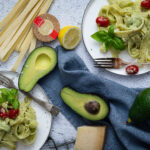 Zwei Teller mit Avocado-Pasta, Kirschtomaten, Basilikum. Nebenbei frische Avocado, Zitronenscheibe, Parmesan und Nudeln.