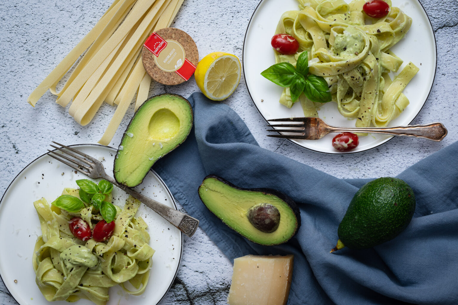 Zwei Teller mit Avocado-Pasta, Kirschtomaten, frischem Basilikum und Parmesan, daneben eine halbierte Avocado.