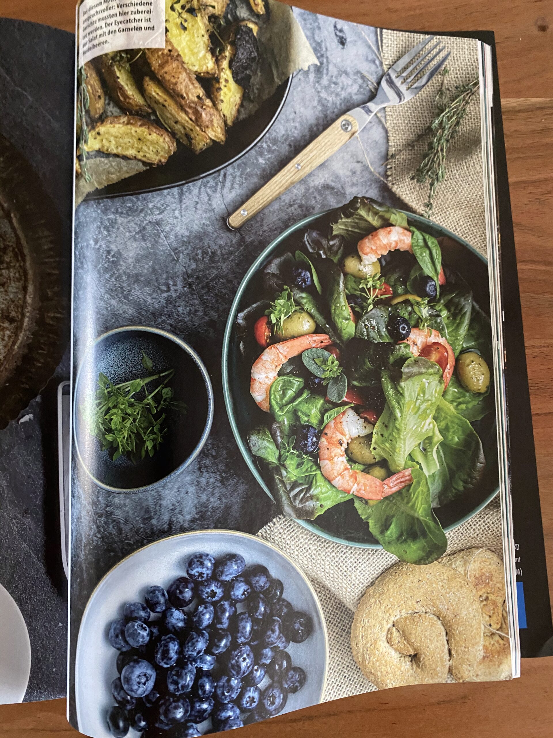 Pictures-Magazin-4 Tisch mit Salat mit Garnelen, Kartoffelecken, Blaubeeren, frischen Kräutern und einem Brötchen auf Schieferplatte.