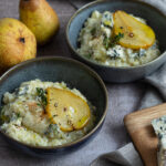 Zwei Schalen Risotto mit Birne und Blauschimmelkäse, daneben zwei ganze Birnen und Käse auf einem Holzbrett.