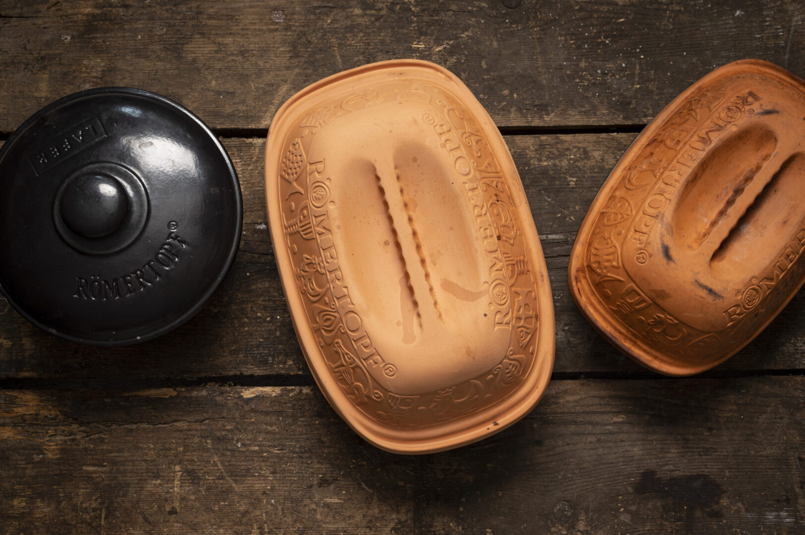 Drei Römertopf-Behälter, zwei braune und einer schwarz, auf einem Holztisch liegend, von oben betrachtet.