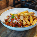 Schale mit Currywurst, Pommes, Ketchup, Zwiebelwürfeln und einem Klecks Mayonnaise auf einem Holztisch.