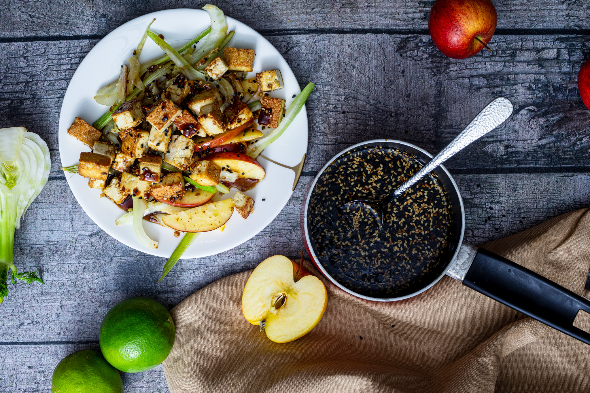 Teriyaki-Sauce-selber-machen_01 Salat mit Tofu und Apfelscheiben auf Teller, daneben Soßenpfanne mit Sesam, Äpfel, Limetten und Fenchel auf Holztisch.