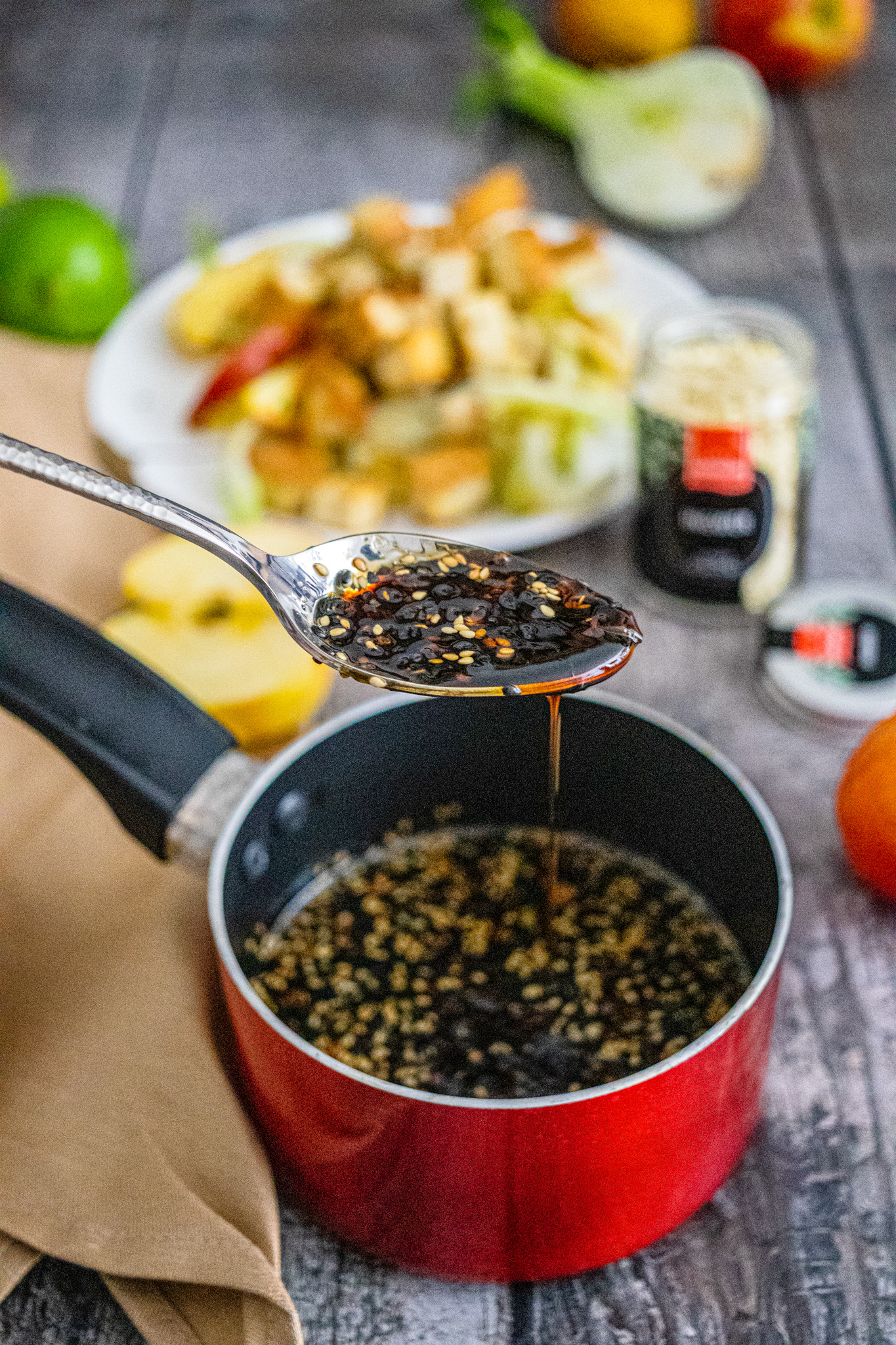 Löffel mit dunkler Sauce über rotem Topf; im Hintergrund unscharfer Salat auf Teller.
