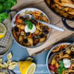 Schalen mit Muschelnudeln, Tomaten, Oliven und Basilikum, garniert mit Mozzarella. Zitrone, Knoblauch und Basilikum im Hintergrund.