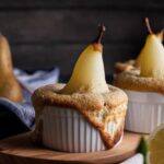 Birnenaufläufe in weißen Förmchen mit karamellisierter Kruste, darauf poelierte Birnenhälften. Im Hintergrund weitere Birnen.