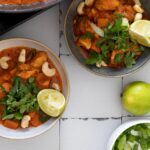 Zwei Schalen mit Curry aus Gemüse, Cashewnüssen und Limettenscheiben, daneben eine ganze Limette und Frühlingszwiebeln.
