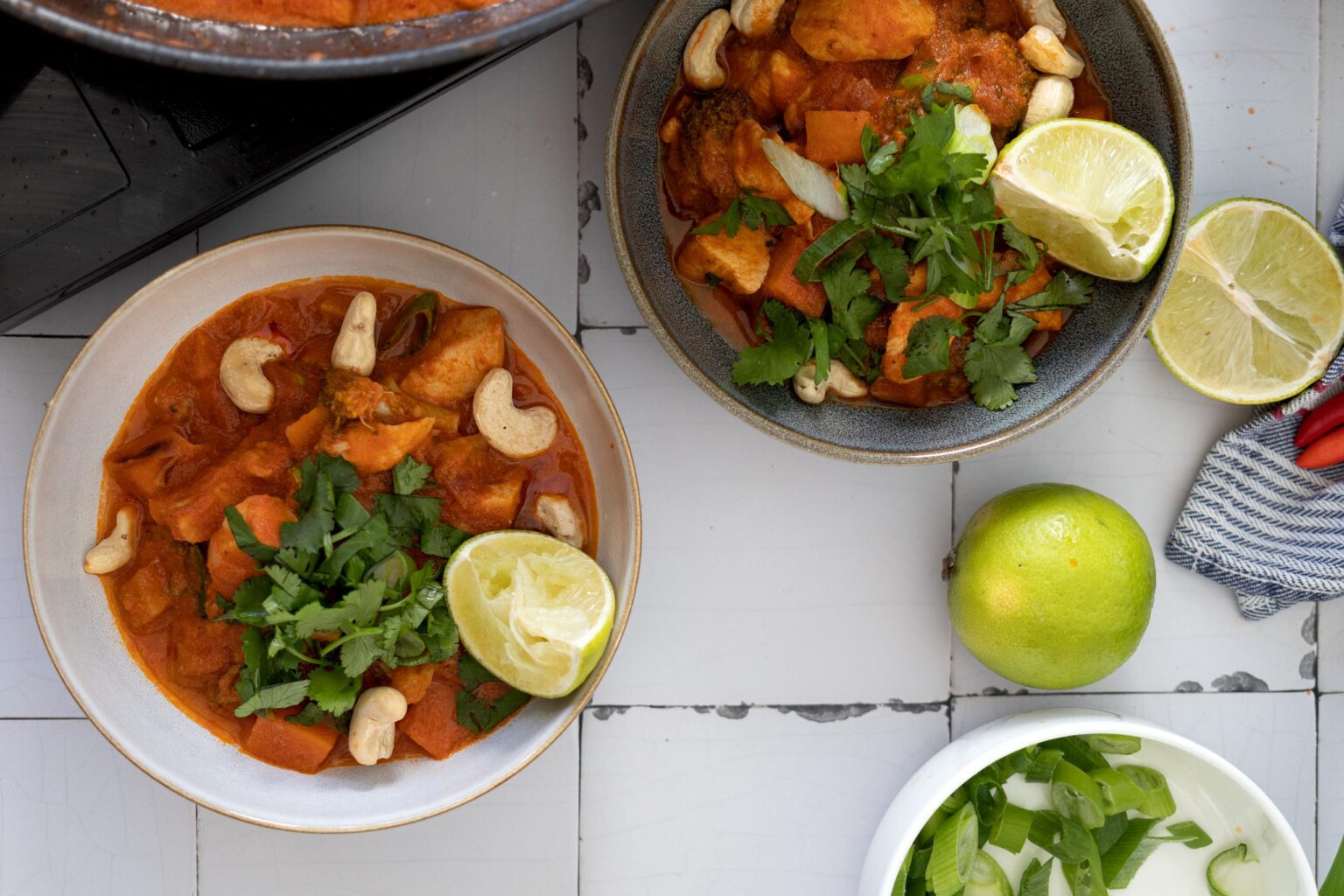 Zwei Schalen mit Curry aus Gemüse, Cashewnüssen und Limettenscheiben, daneben eine ganze Limette und Frühlingszwiebeln.